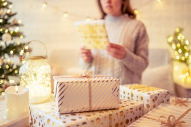 Noel ağacının yanındaki koltukta oturan ve el yapımı hediye kutularını masaya koyan kadın. Aile için hediyeler.