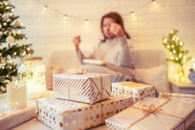 Noel ağacının yanındaki koltukta oturan ve el yapımı hediye kutularını masaya koyan kadın. Aile için hediyeler.