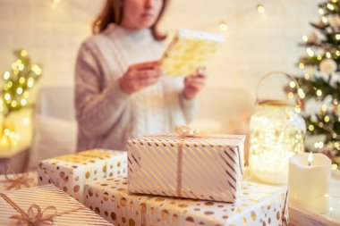 Noel ağacının yanındaki koltukta oturan ve el yapımı hediye kutularını masaya koyan kadın. Aile için hediyeler.