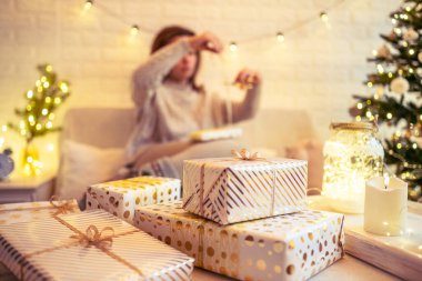Noel ağacının yanındaki koltukta oturan ve el yapımı hediye kutularını masaya koyan kadın. Aile için hediyeler.