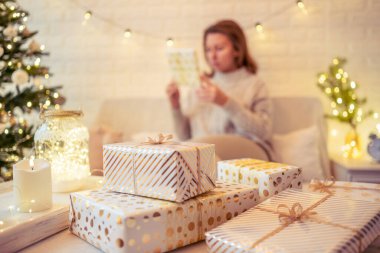 Noel ağacının yanındaki koltukta oturan ve el yapımı hediye kutularını masaya koyan kadın. Aile için hediyeler.