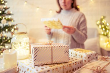Noel ağacının yanındaki koltukta oturan ve el yapımı hediye kutularını masaya koyan kadın. Aile için hediyeler.