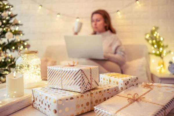クリスマスツリーの近くに座ってギフトのアイデアを検索し テーブルの上にそれを置く手作りのギフトボックスを梱包するためにラップトップを使用してソファの上に焦点を当てていない ぼやけた女性 家族へのプレゼント — ストック写真