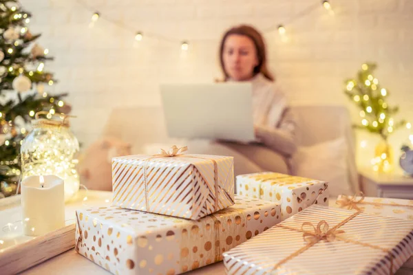 クリスマスツリーの近くに座ってギフトのアイデアを検索し テーブルの上にそれを置く手作りのギフトボックスを梱包するためにラップトップを使用してソファの上に焦点を当てていない ぼやけた女性 家族へのプレゼント — ストック写真