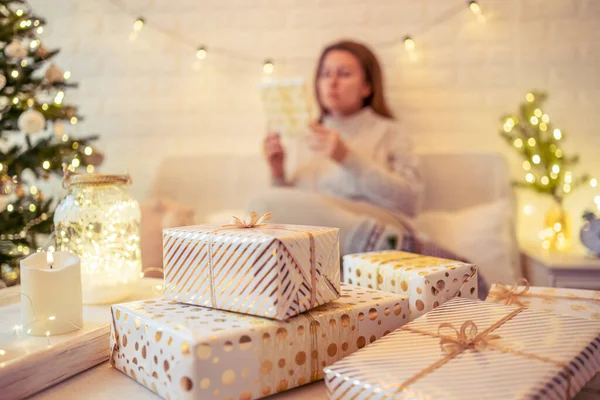 Noel ağacının yanındaki koltukta oturan ve el yapımı hediye kutularını masaya koyan kadın. Aile için hediyeler.