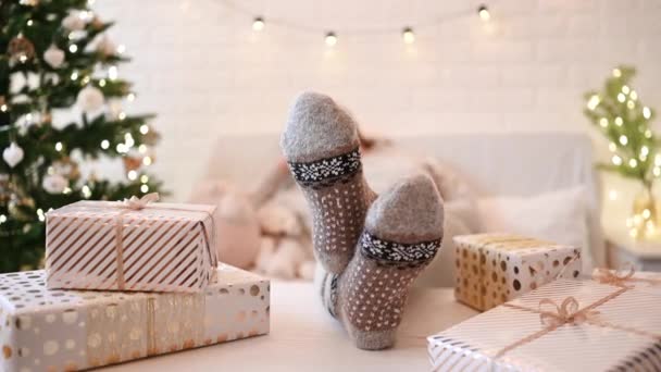 クリスマスツリーの近くのソファに座っている女性 女性の足 冬の足は テーブルの上に置き 手作りのギフトボックスを梱包した後 リラックスしたウールの靴下 家族へのプレゼント — ストック動画