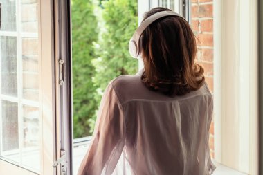 Kulaklıklı genç bir kadın köy evinin banyo penceresinden müzik dinliyor ve yaz sabahının erken saatlerinde temiz hava soluyor. Arka plan.
