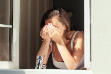 Genç bir esmer kadın güneşli bir sabahın erken saatlerinde yüzünü banyoda yıkar. Bir kır evinin açık penceresinden bak 
