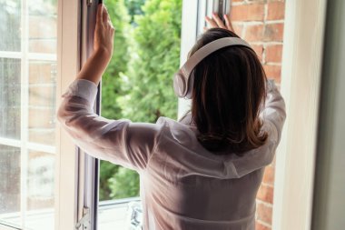 Kulaklıklı, müzik dinleyen genç, güzel, mutlu bir kadın köy evinin penceresini açıyor ve yaz sabahının erken saatlerinde temiz hava soluyor. Arka plan.