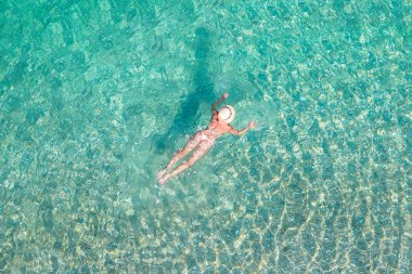 Top, aerial view. Young beautiful woman in a hat and white bikini swimming in sea water on the sand beach. Drone, copter photo. Summer vacation. View from above. 