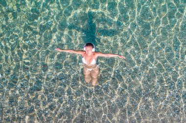 Top, aerial view. Athletic body of young beautiful woman in headphones listening music lying and swimming in white bikini in sea water on the sand beach. Drone, copter photo. Summer vacation.