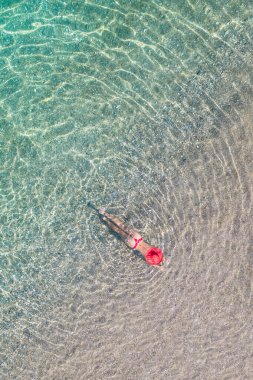 Üst Manzara. Kırmızı şapkalı ve bikinili genç bir kadın kumsalda deniz suyunda güneşleniyor. Drone, helikopter fotoğrafı. Yaz tatili. Yukarıdan görüntüle. 