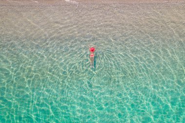 Üst Manzara. Kırmızı şapkalı ve bikinili genç bir kadın kumsalda deniz suyunda güneşleniyor. Drone, helikopter fotoğrafı. Yaz tatili. Yukarıdan görüntüle. 