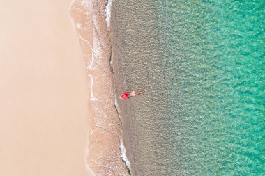 Üst Manzara. Genç, çıplak, kırmızı şapkalı bir kadın kumsalda deniz suyunda güneşleniyor. Drone, helikopter fotoğrafı. Yaz tatili. Yukarıdan görüntüle. 