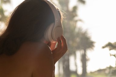 Güneş yatağında terasta uzanmış, gün batımında palmiye ağaçlarına bakan kulaklıklı kadın silueti. Kadın dinlendirici müzik. Arka plan.
