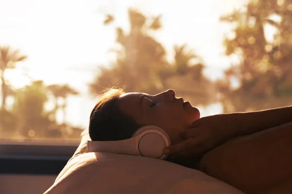 一个女人的轮廓 带着耳机躺在枕头上 在卧室对面的全景窗户上 日落时可以看到棕榈树海滩 女性放松听音乐 — 图库照片