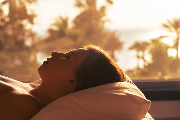 女性のシルエットはベッドの上に横たわっており 夕日でヤシの木の海のビーチの景色を望むパノラマの窓の反対側の寝室で枕の上に寝て — ストック写真