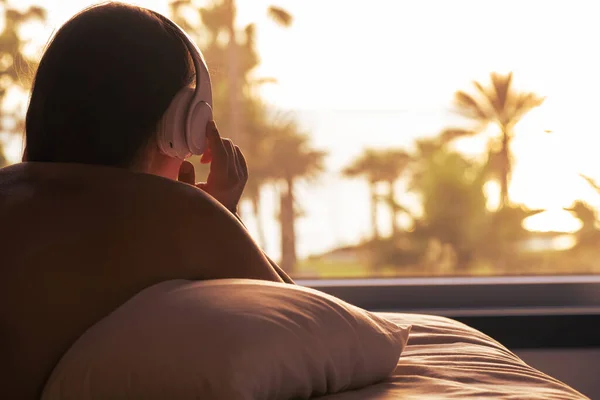 一个女人的轮廓 带着耳机躺在枕头上 在卧室对面的全景窗户上 日落时可以看到棕榈树海滩 女性放松听音乐 — 图库照片