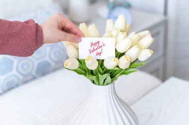 Masanın üzerinde beyaz bir vazoda duran beyaz lale buketinden Sevgililer Günü kartı çeken bir kadın.. 