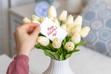 Masanın üzerinde beyaz bir vazoda duran beyaz lale buketinden Sevgililer Günü kartı çeken bir kadın.. 