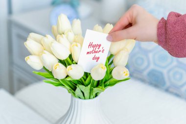 Masadaki beyaz vazodaki beyaz lale buketinden mutlu anneler günü sözleriyle tebrik kartı çeken bir kadın.. 