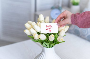 Masadaki beyaz vazodaki beyaz lale buketinden mutlu anneler günü sözleriyle tebrik kartı çeken bir kadın.. 