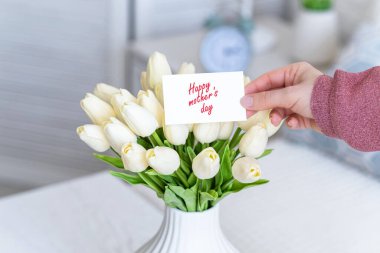 Masadaki beyaz vazodaki beyaz lale buketinden mutlu anneler günü sözleriyle tebrik kartı çeken bir kadın.. 