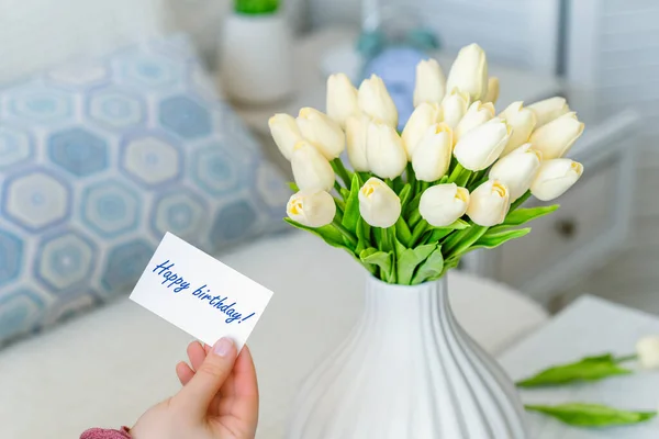 女はテーブルの上に白い花瓶に白いチューリップの花の花束から幸せな誕生日の言葉でグリーティングカードを引く — ストック写真