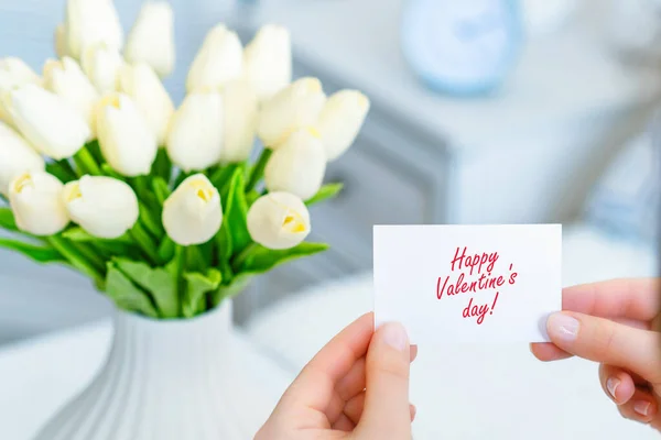 幸せなバレンタインデーの言葉で挨拶カードを引く女性白いチューリップの花束からテーブルの上に白い花瓶に花 — ストック写真