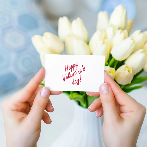 幸せなバレンタインデーの言葉で挨拶カードを引く女性白いチューリップの花束からテーブルの上に白い花瓶に花 — ストック写真