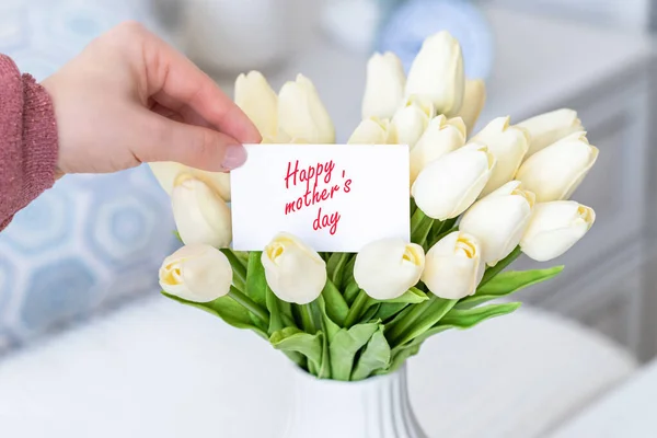 女が挨拶カードを引く幸せな母の日の言葉から花束の白いチューリップの花でテーブルの上に白い花瓶 — ストック写真