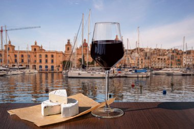 Bir bardak kırmızı şarap, Brie peyniri ve liman manzaralı tekne ve Malta 'nın tarihi şehir merkezi Birgu..