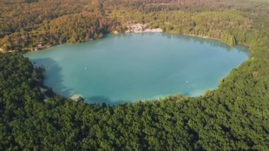Aerial top-down flight over the amazing small lake of a perfectly round shape. Cloudy sky reflected in the clear turquoise water of a pond surrounded by trees and plants. 