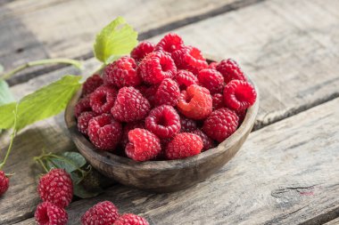 Tahta arka planda yeşil ahududu yapraklarıyla çevrili bir kasede sulu olgun ahududular. Boşluğu kopyala