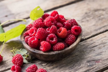 Tahta arka planda yeşil ahududu yapraklarıyla çevrili bir kasede sulu olgun ahududular. Boşluğu kopyala