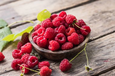 Tahta arka planda yeşil ahududu yapraklarıyla çevrili bir kasede sulu olgun ahududular. Boşluğu kopyala