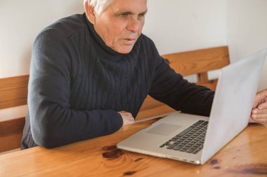 Gülümseyen yaşlı adam kameraya el sallıyor dizüstü bilgisayarında video görüşmesi yapıyor, mutlu yaşlı erkek evde oturup modern teknolojileri ve kablosuz bağlantıyı kullanıyor.