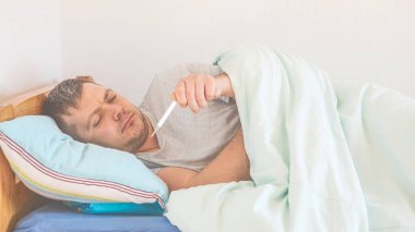 Evdeki yatakta termometresi olan hasta bir adam. Hastalık kavramı, ateş belirtileri.