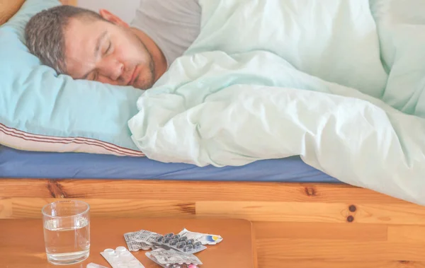 Sick man sleeping in a bed, pills and water glass in the foreground. Concept of illness, fever, coronavirus symptoms