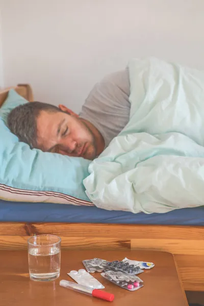 Sick man sleeping in a bed, pills and water glass in the foreground. Concept of illness, fever, coronavirus symptoms