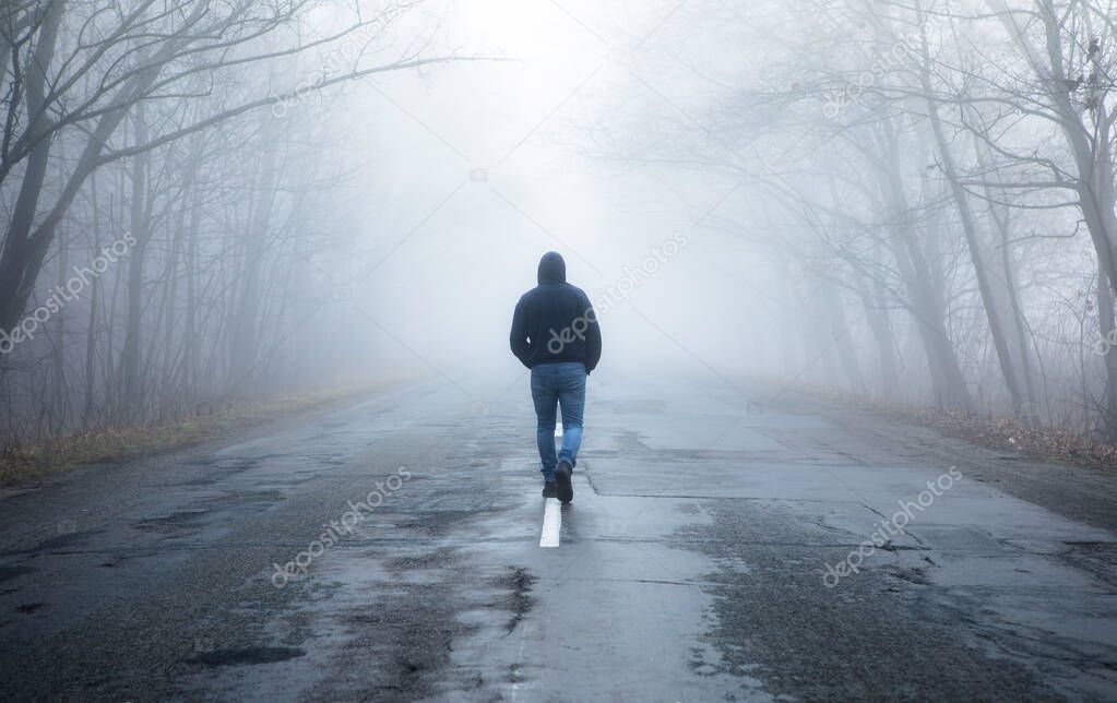 Lonely man walking in fog away road. Rural landscape with road in morning mist. Warm autumn colors. Dark mysterious background