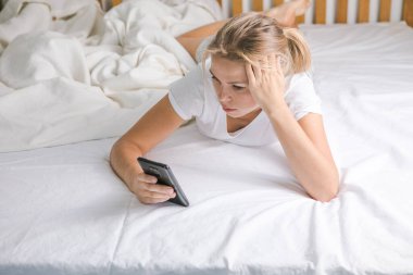 Cep telefonuyla yatakta uzanan gülümseyen orta yaşlı bir kızın portresi. Evde cep telefonu kullanan mutlu sarışın kadın. Güzel genç bir bayan yatak odasında akıllı telefonuyla yazı yazıyor.