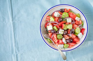 Beyaz peynirli Yunan salatası, zeytin, domates, salatalık ve kırmızı soğan sağlıklı vejetaryen Akdeniz diyeti yemekleri, düşük kalorili yiyecekler. Mavi kumaş arkaplan, üst görünüm
