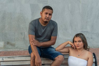 Genç bir çift parkta bankta oturuyor ve kameraya bakıyor..