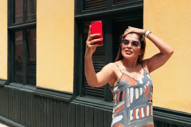 Uzun saçlı ve gözlüklü İspanyol kadın şehir sokaklarında yürüyor ve cep telefonuyla selfie çekiyor..