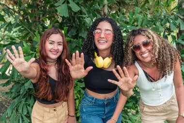 Üç arkadaş dışarıda kameraya bakıyor ve ellerini gösteriyorlar..