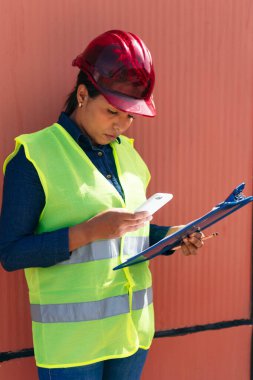 Koruyucu yelekli genç bir kadın bir fabrikada cep telefonunu kontrol ediyor.