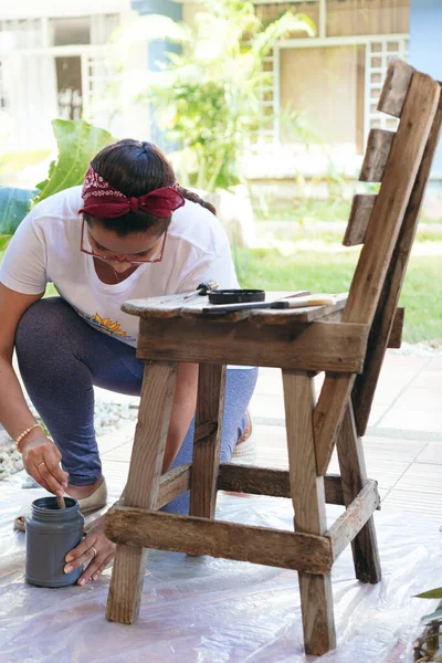 Arka bahçede genç bir kadın elinde bir bant tutarken restorasyon için bir sandalye hazırlıyor.
