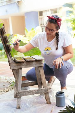 Bahçedeki sandalyeyi boyamak ve restore etmek için öne eğilen genç bir kadın.