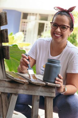 Arka bahçedeki kadın eski bir sandalye boyuyor ve gülümsüyor.